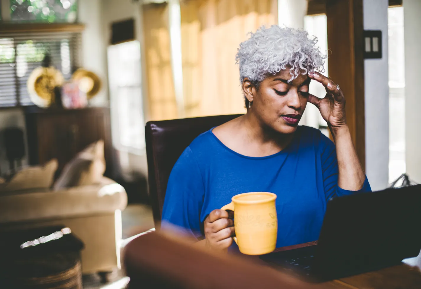 A woman rubs her forehead while holding a coffee mug and using a laptop.