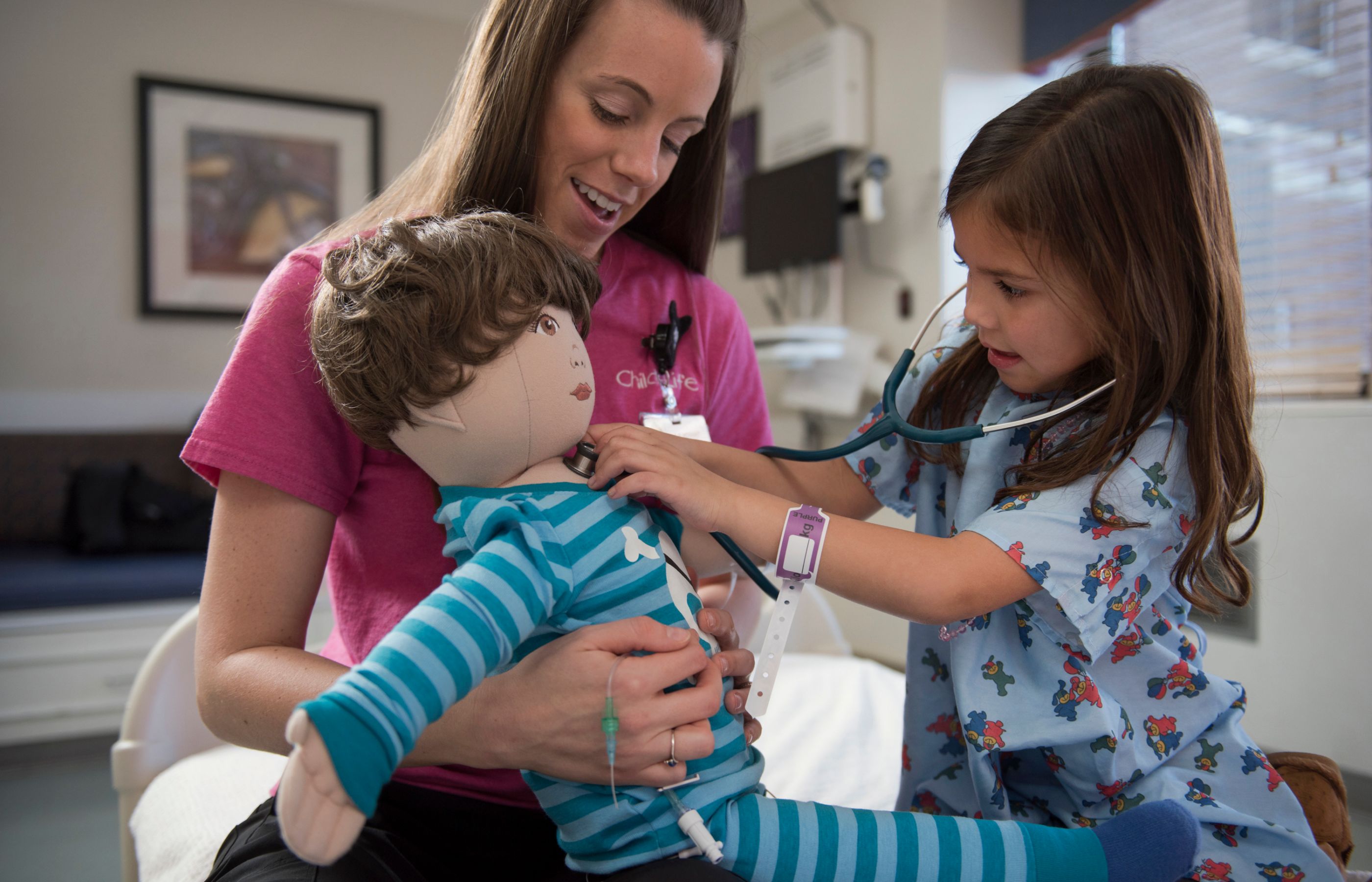 A nurse is holding a teddy bear as a young child is using the stethoscope to listen to the bears heartbeat.