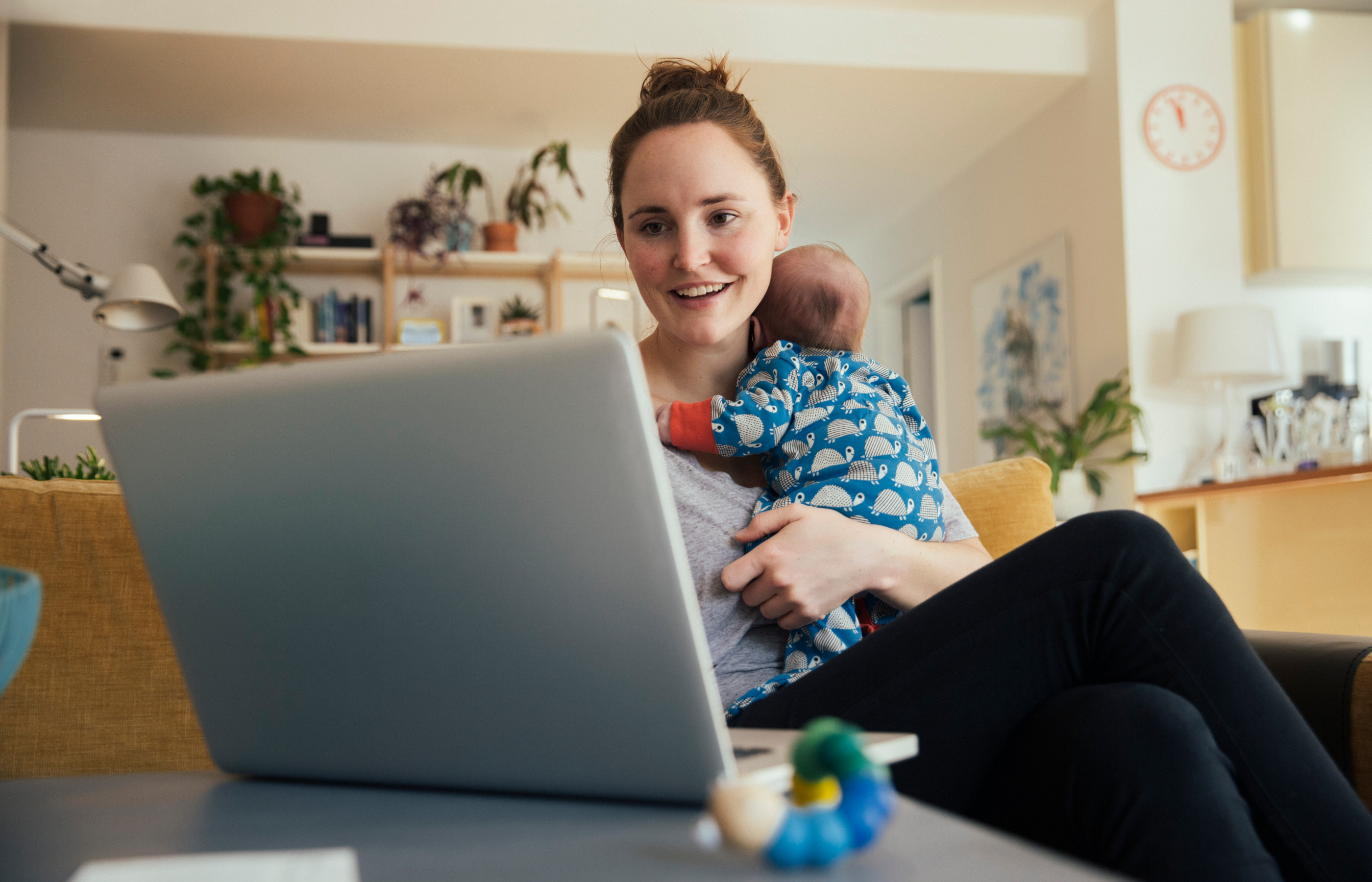 A mother is sitting on her couch holding her baby while looking at a laptop. 