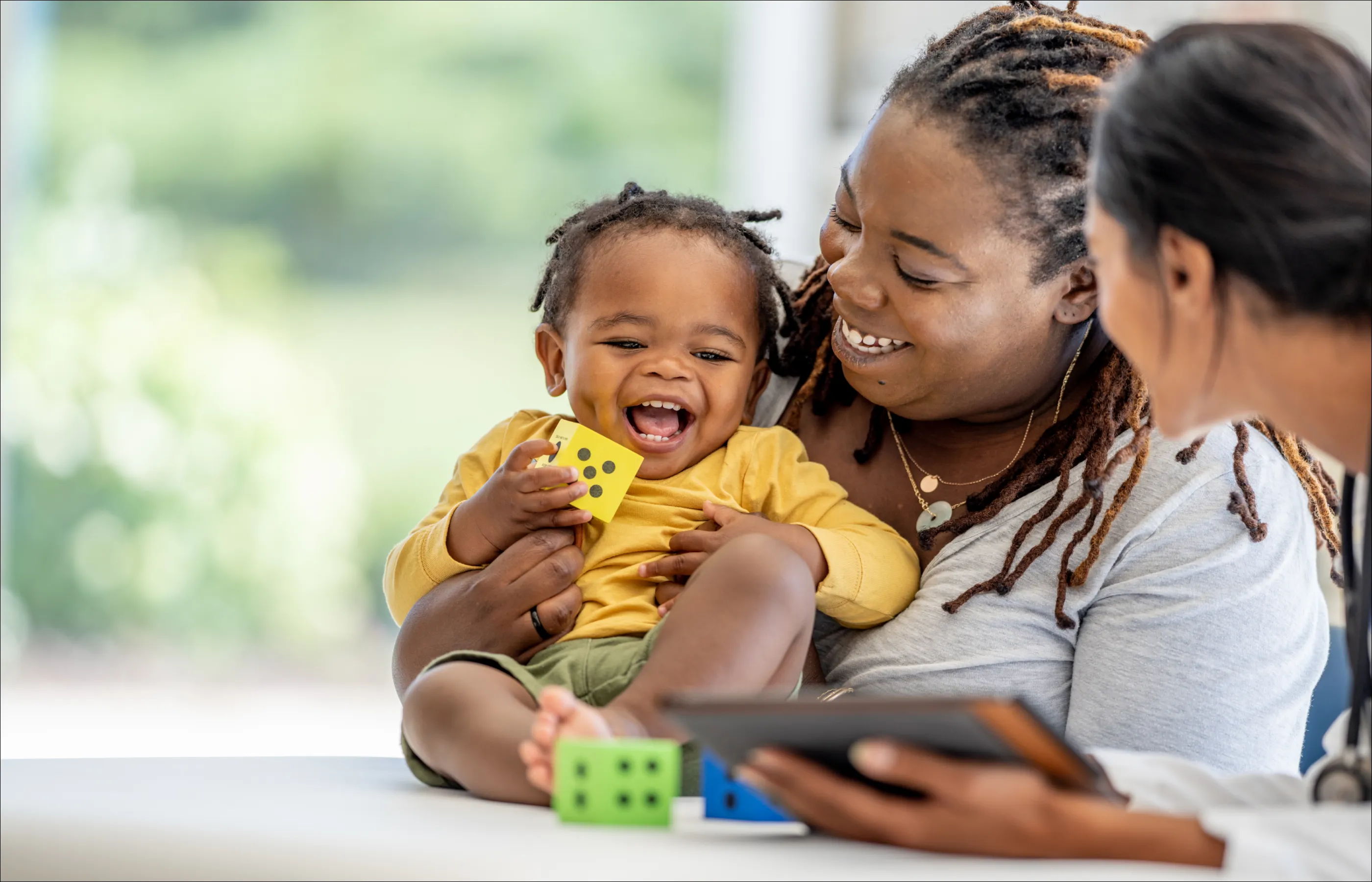 A mom, her toddler, and a provider holding a tablet smile as the baby plays with dice blocks