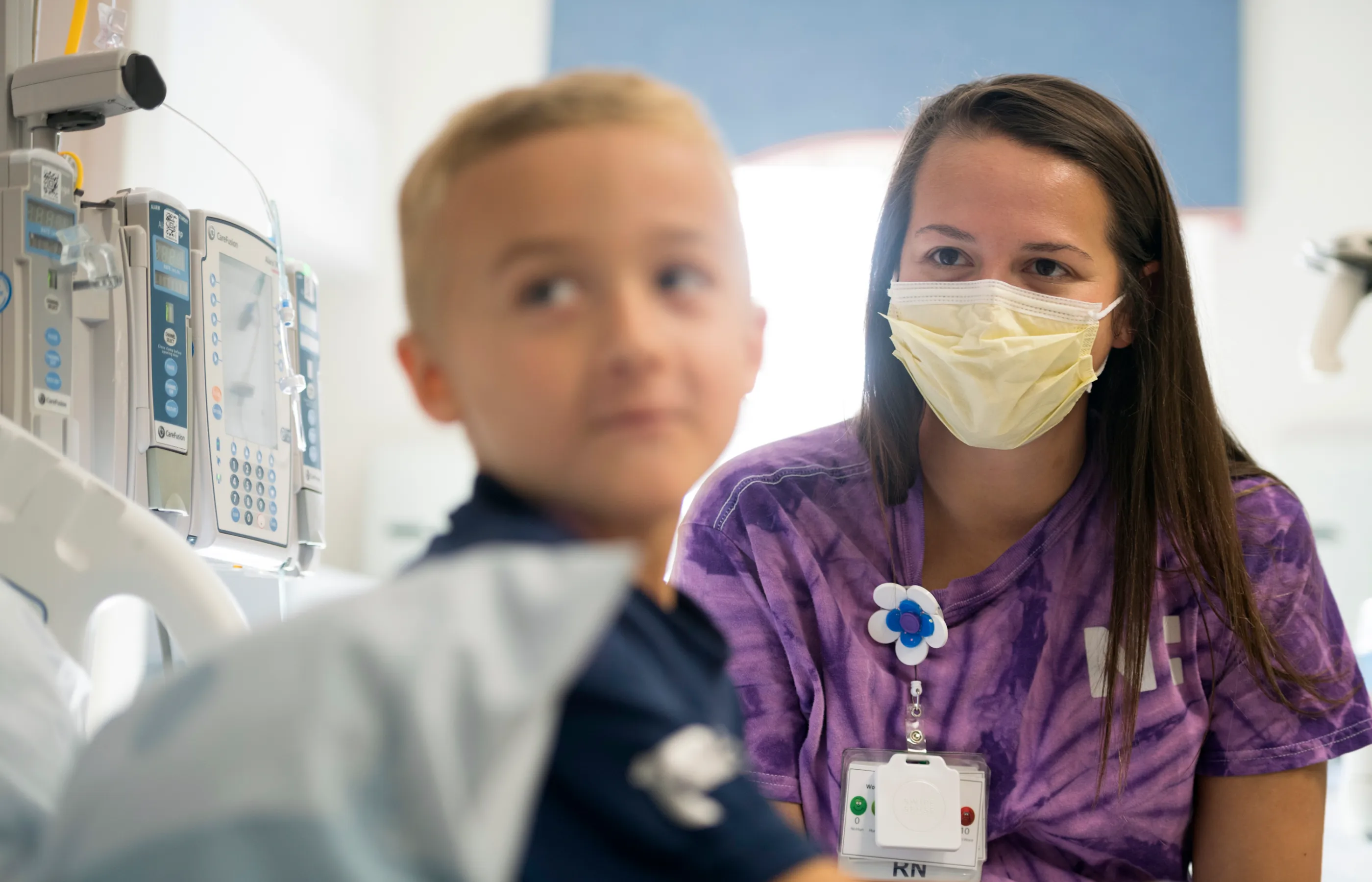 A masked RN smiles at a young boy in a pediatric hospital room
