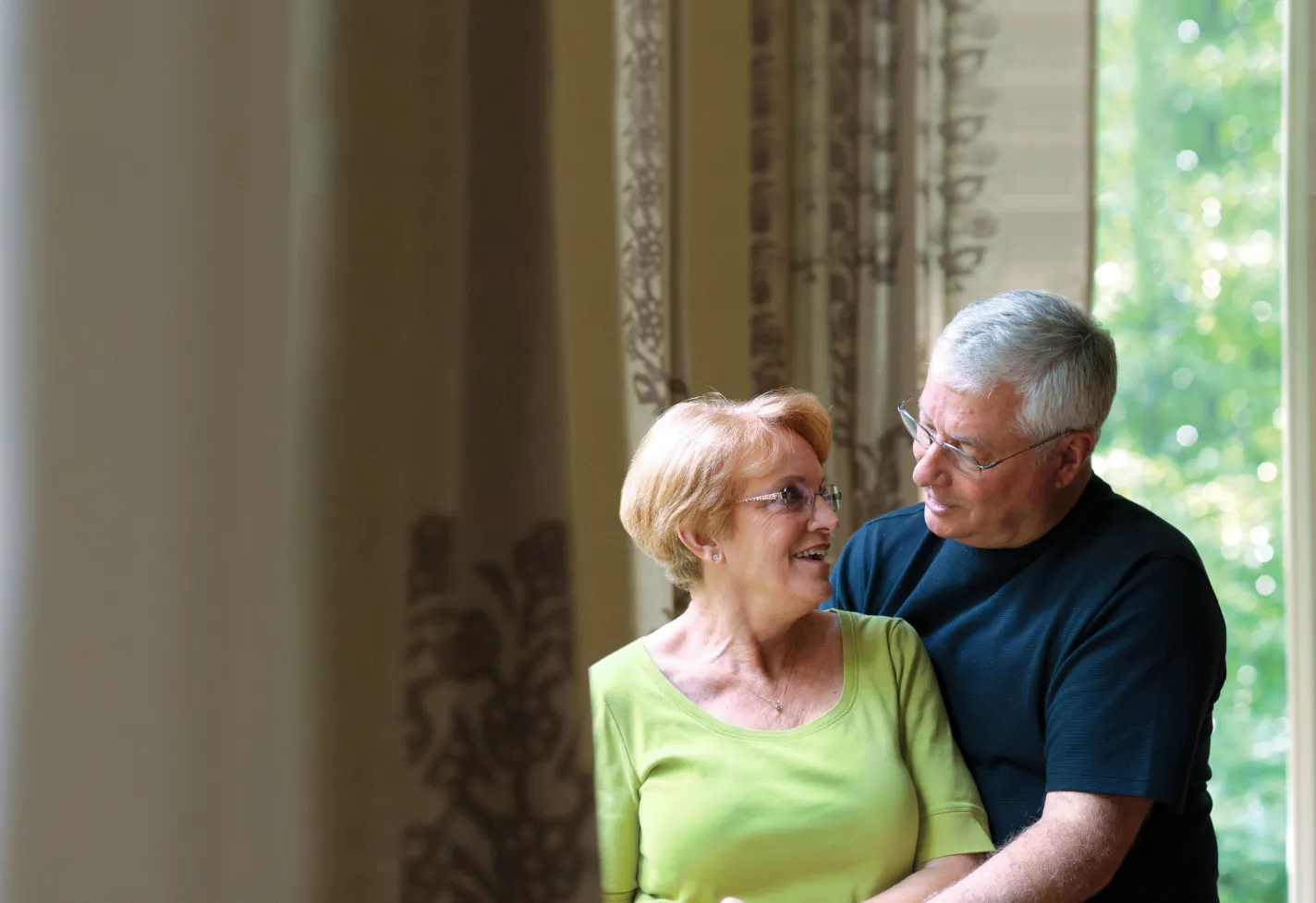 A senior man is standing behind his partner as he holds her hand. 