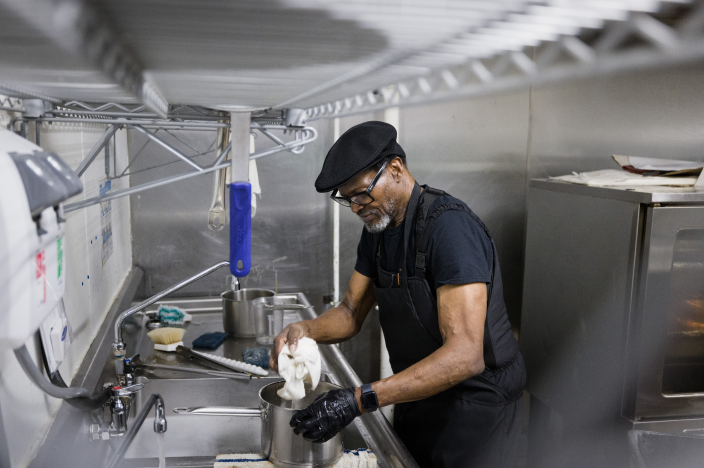 A man wearing a hat and gloves is working in a kitchen, handling a cloth near a pot in a stainless steel sink.