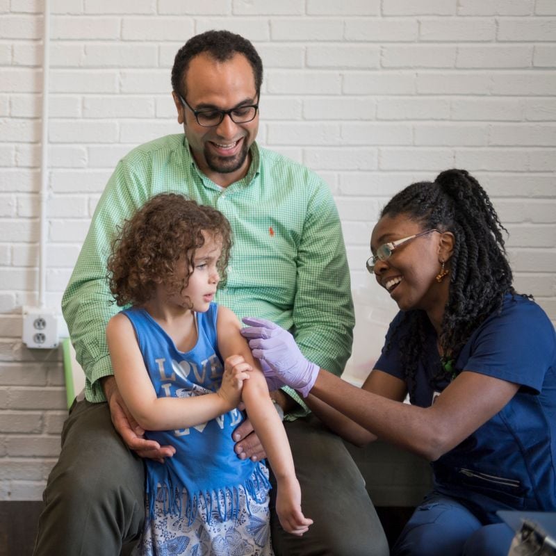 A Novant Health nurse, is administering a vaccine to a child who is accompanied by their dad.