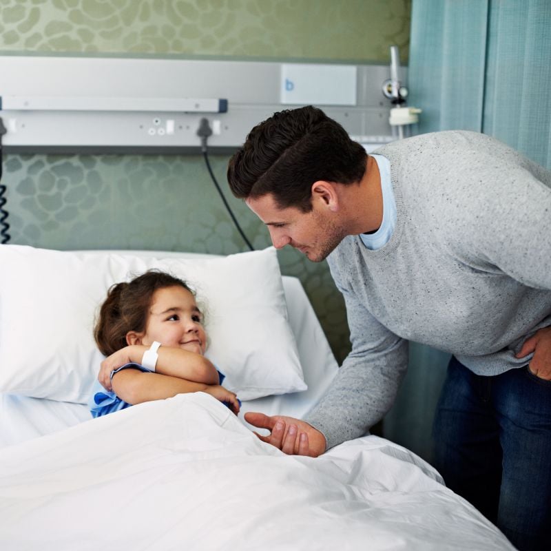A father is comforter his daughter as she rests in a hospital bed. 