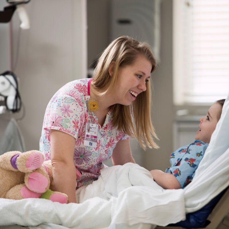 An RN sits on a hospital bed and laughs with a little boy and his teddy bear