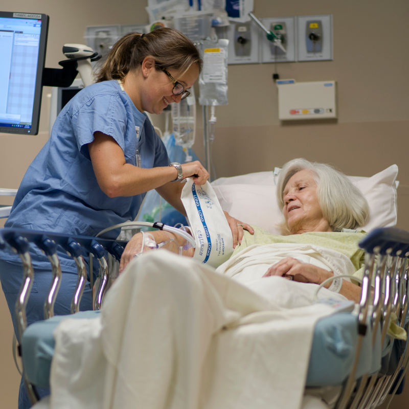 Nurse and patient are in the post-op area of the hospital. The patient is lying in bed, while the nurse wraps a blood pressure cuff around her arm.  